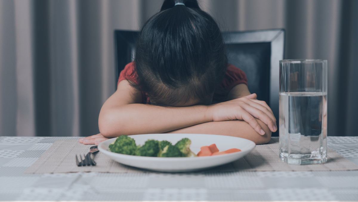 A child lays her head in her arms on her table with a plate of uneaten food in front of her.
